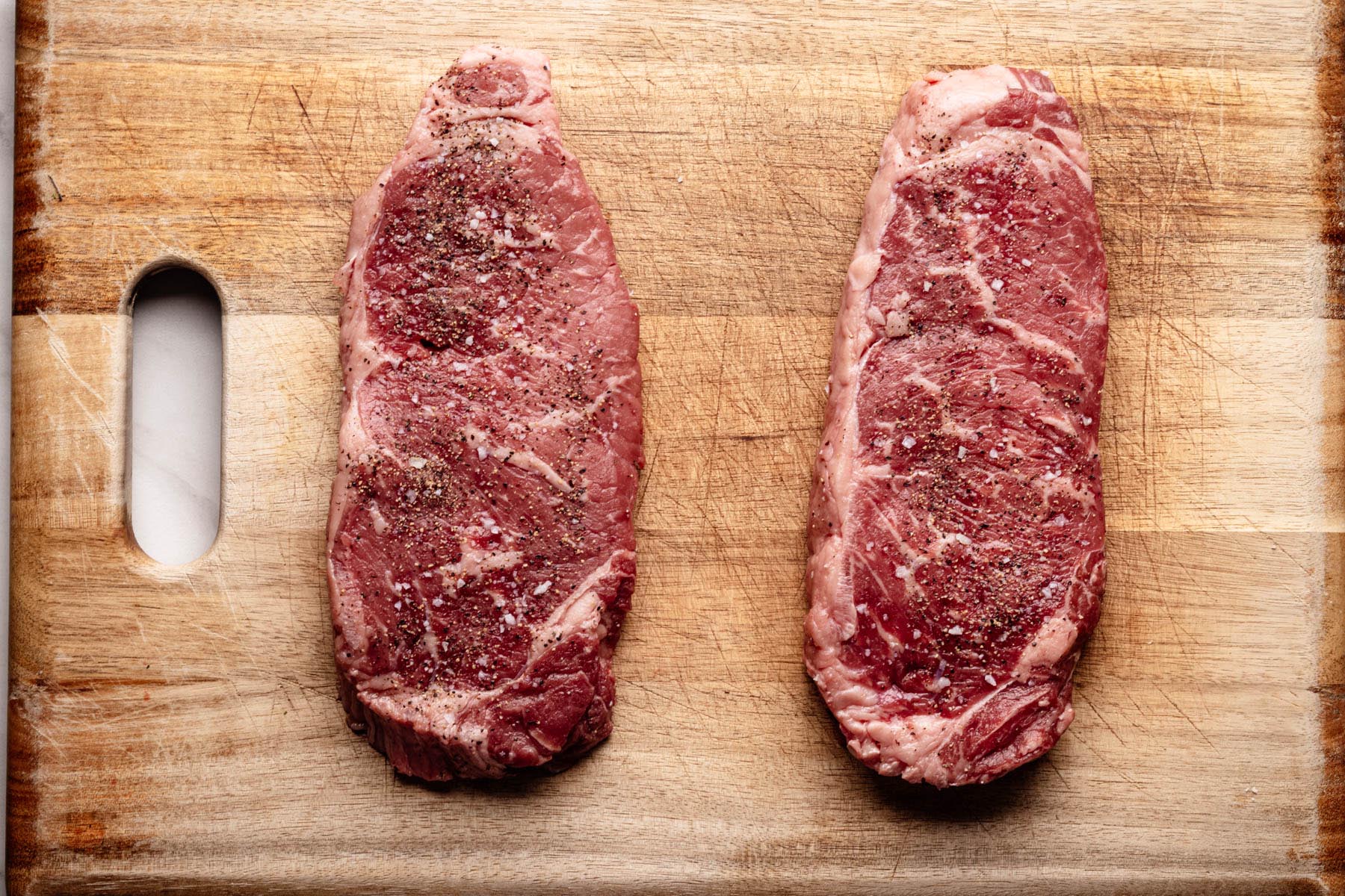 Two raw strip steaks seasoned with salt and black pepper on a wooden cutting board.