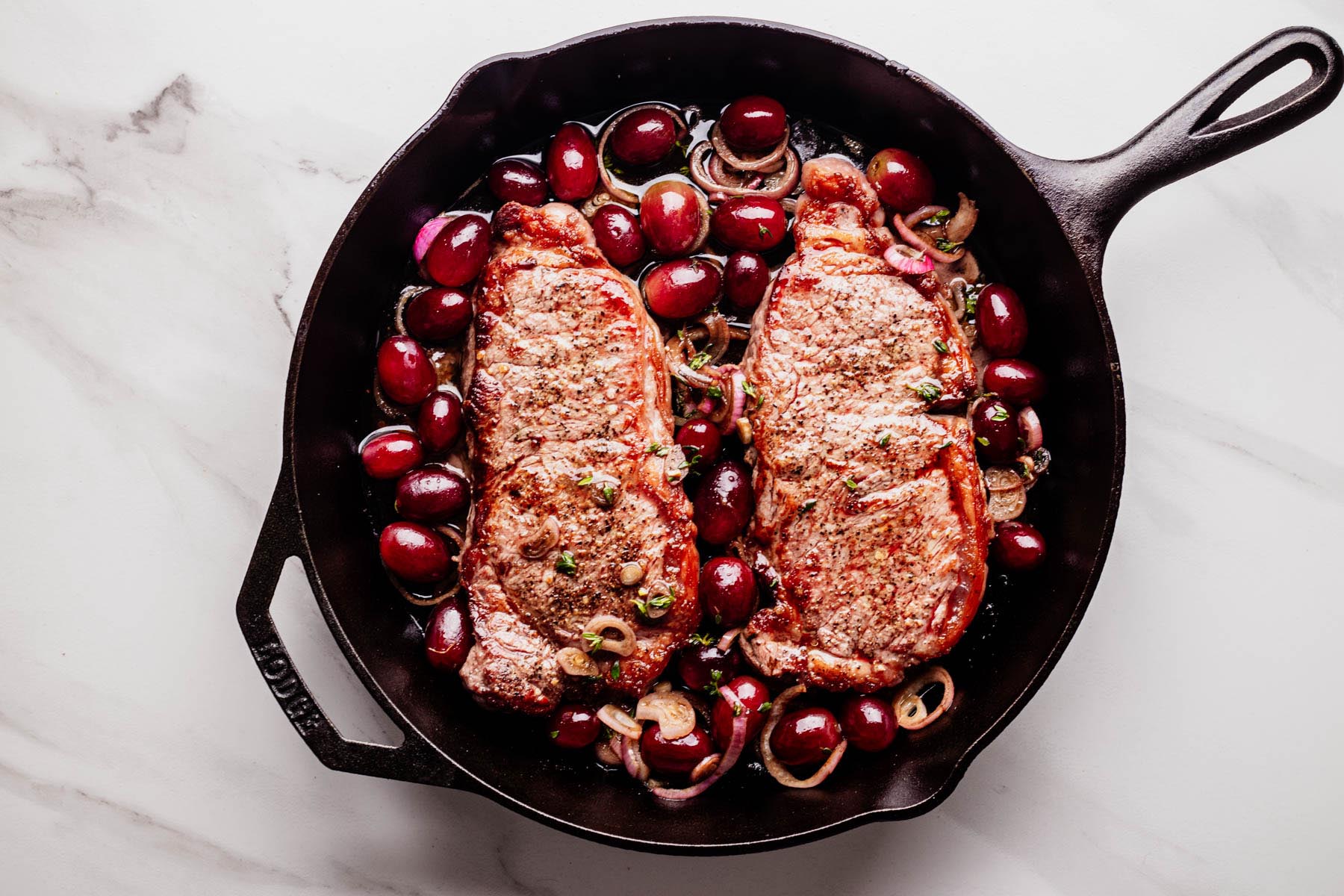 Two seared steaks in a cast iron skillet with roasted red grapes, sliced shallots, and fresh thyme, ready to transfer to the oven.