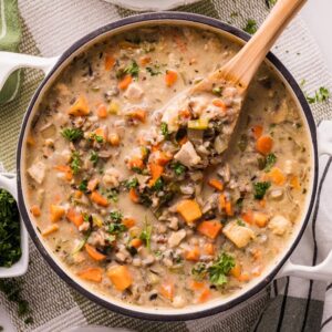 Overhead of a Dutch oven filled with creamy chicken and wild rice soup—carrots, celery, mushrooms, and parsley—with a wooden spoon lifting a scoop.