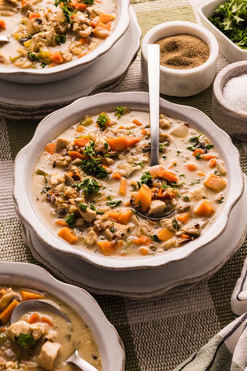 White bowl of creamy chicken and wild rice soup with carrots, sweet potato, mushrooms, and parsley, a spoon resting inside, on a green plaid cloth.
