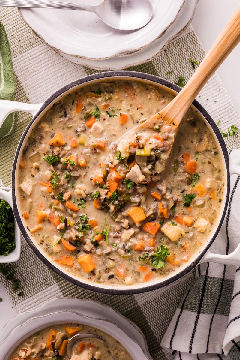 Overhead view of a Dutch oven filled with creamy chicken and wild rice soup&mdash;carrots, celery, mushrooms, and parsley visible&mdash;wooden spoon lifting a hearty scoop beside stacked bowls and a striped towel.