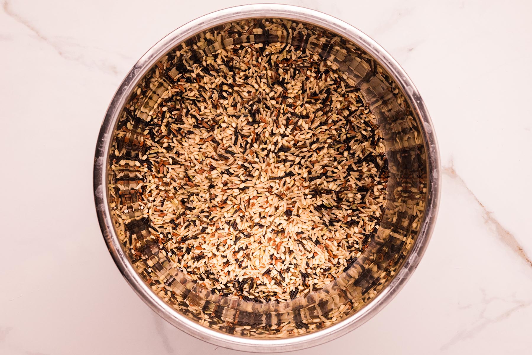 Stainless steel pot filled with rinsed wild rice blend on a white marble counter.