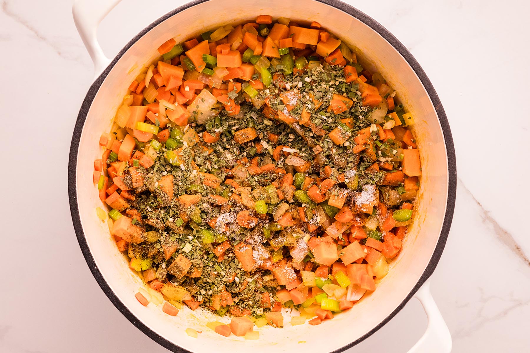 Saut&eacute;ed vegetables in a Dutch oven sprinkled with poultry seasoning, salt, and pepper before stirring. Saut&eacute;ed carrots, celery, onion, and sweet potato in a white Dutch oven, topped with poultry seasoning, salt, and pepper, ready to stir.