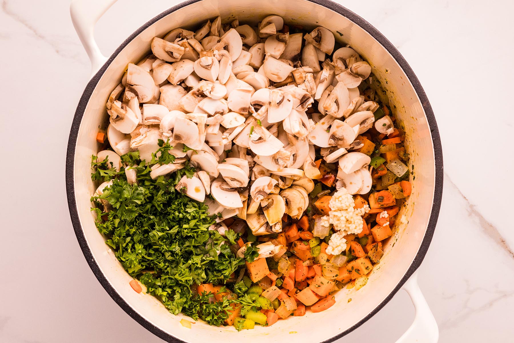 Overhead view of a Dutch oven with the saut&eacute;ed vegetable base topped with sliced mushrooms, chopped parsley, and minced garlic, ready to stir for chicken and wild rice soup.