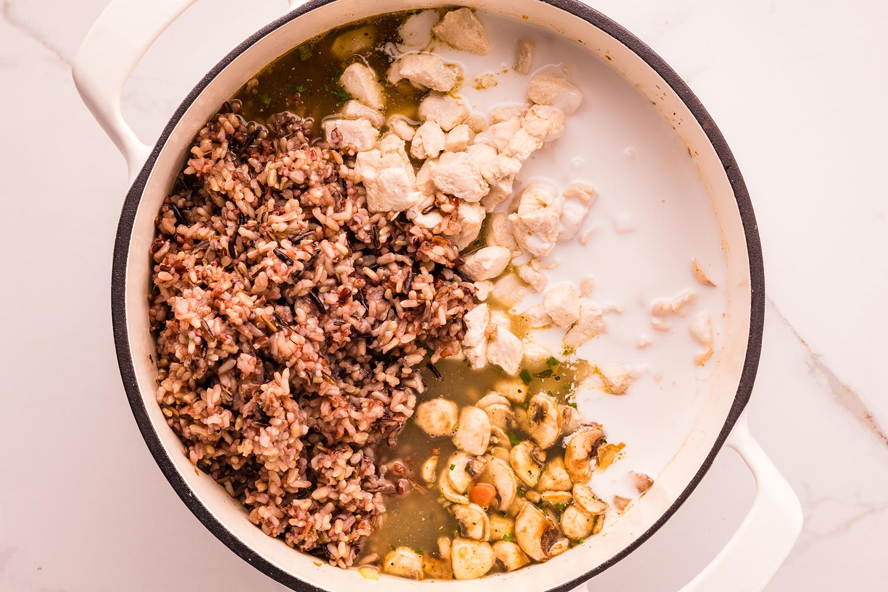 Overhead view of a white Dutch oven with cooked wild rice, seared chicken pieces, saut&eacute;ed mushrooms, broth, and coconut milk just added, ready to stir into creamy chicken and wild rice soup.