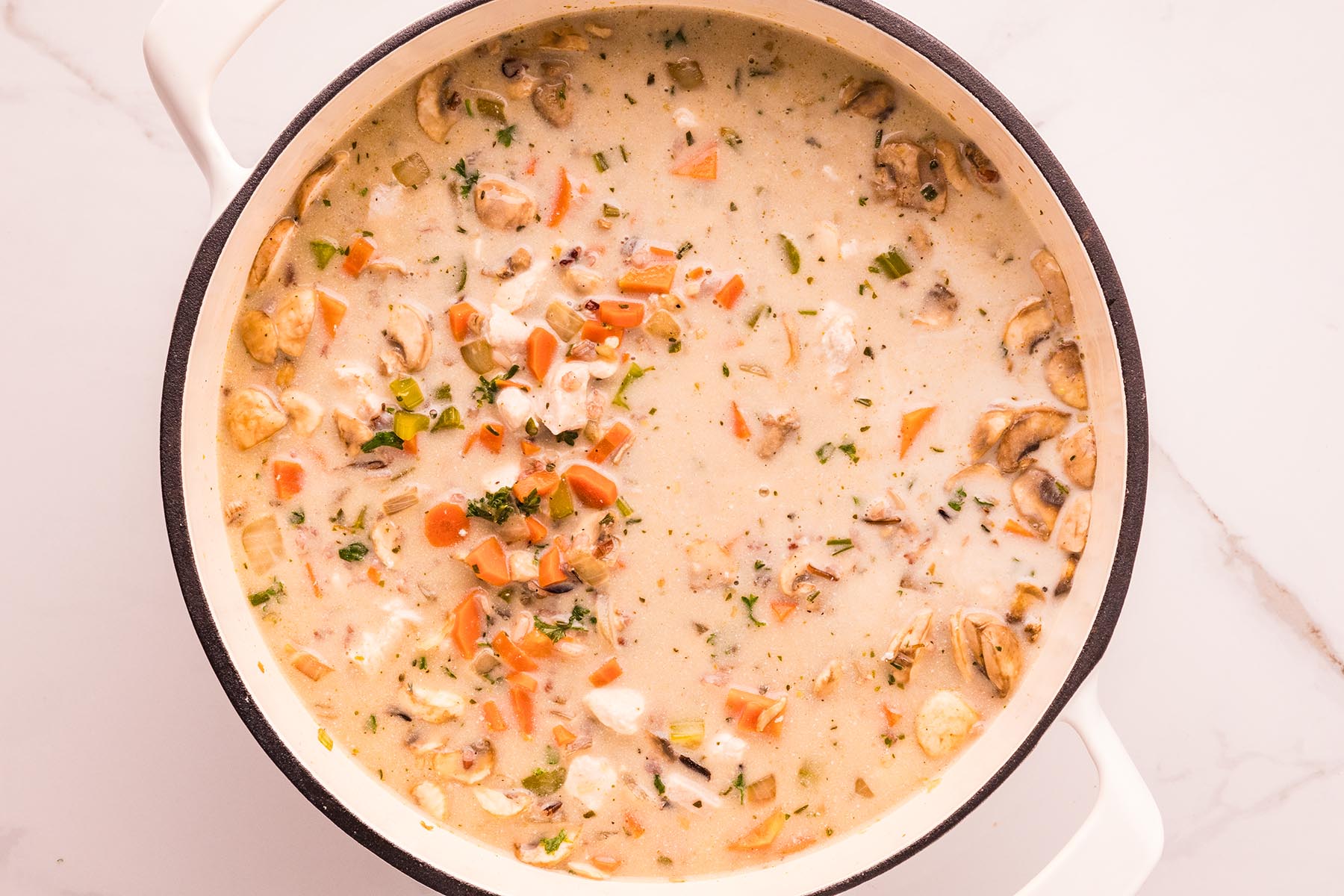 Overhead view of a white Dutch oven filled with creamy chicken and wild rice soup&mdash;tan broth speckled with wild rice, carrots, celery, mushrooms, and parsley, gently simmering.