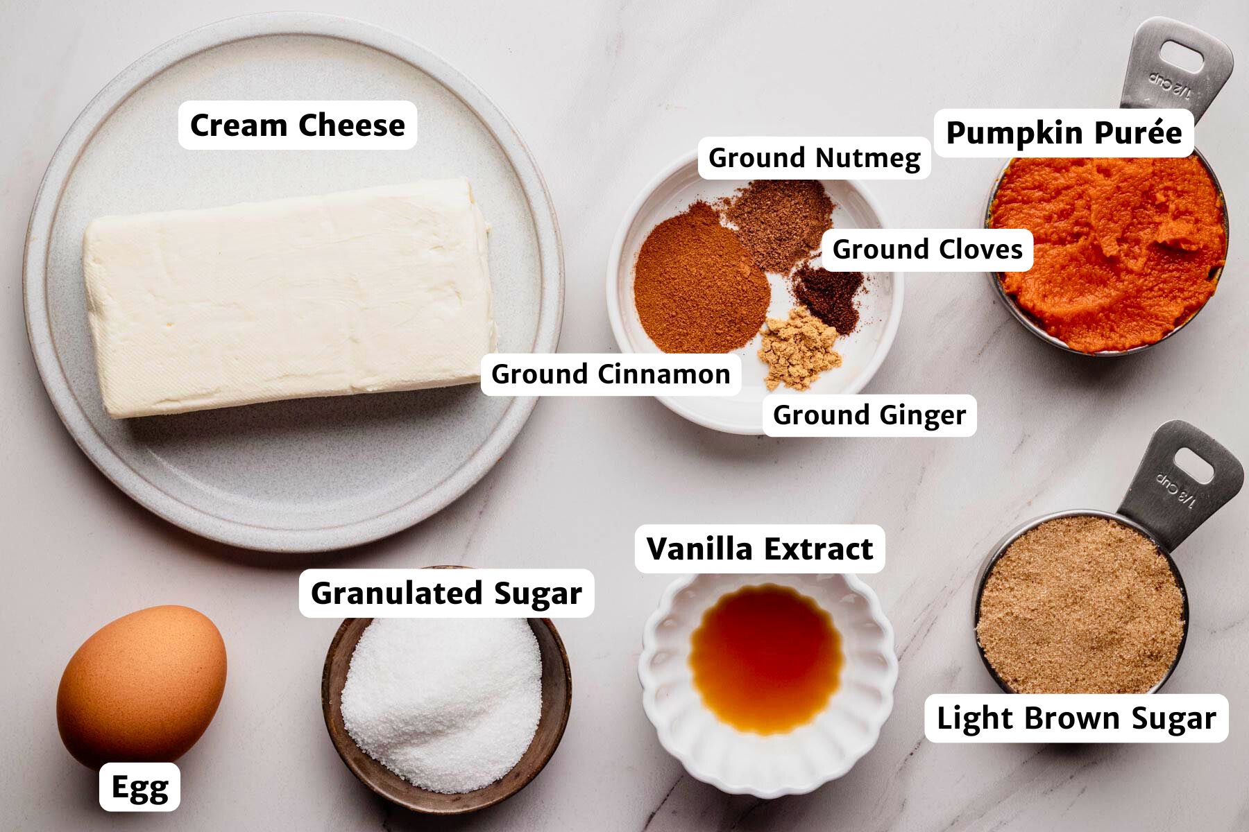 Overhead view of labeled filling ingredients for mini pumpkin cheesecakes, including cream cheese, pumpkin purée, granulated sugar, light brown sugar, egg, vanilla extract, ground cinnamon, ground nutmeg, ground cloves, and ground ginger arranged on a white surface.