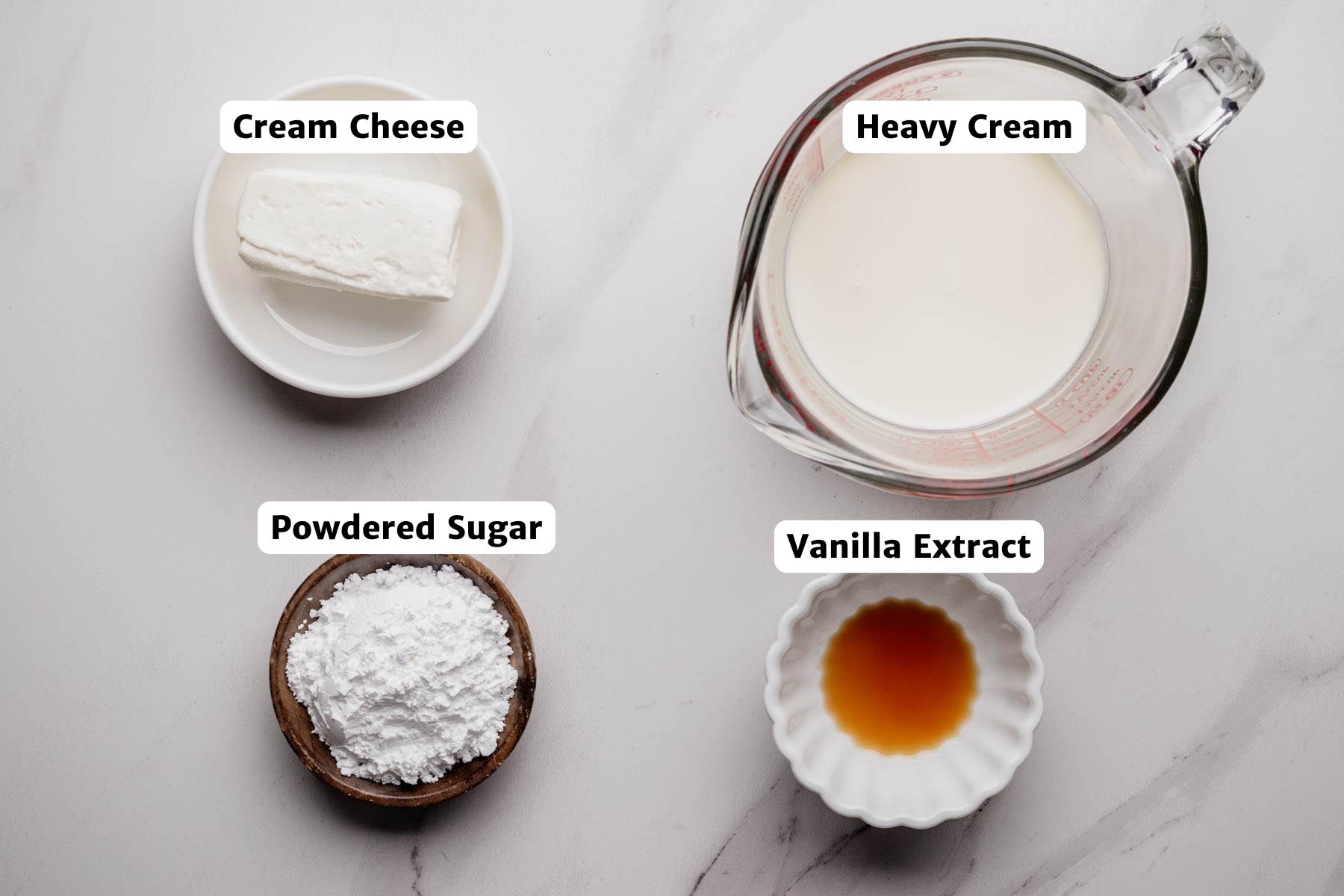 Overhead view of labeled topping ingredients for mini pumpkin cheesecakes, including cream cheese, heavy cream, powdered sugar, and vanilla extract arranged on a white surface.
