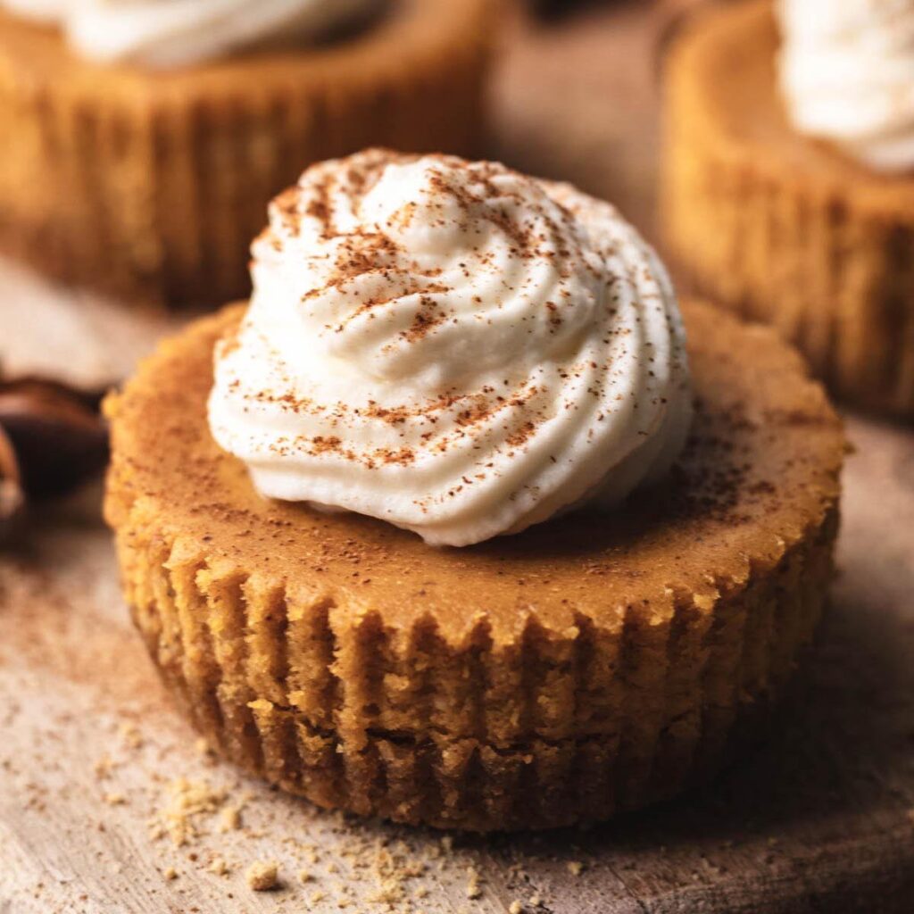 Close-up of a mini pumpkin cheesecake topped with a swirl of whipped cream and a dusting of cinnamon, set on a wooden surface.