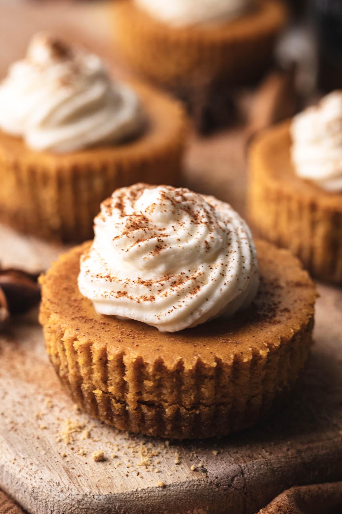 Close-up of a mini pumpkin cheesecake with graham cracker crust, topped with a swirl of cream cheese whipped cream and dusted with cinnamon.