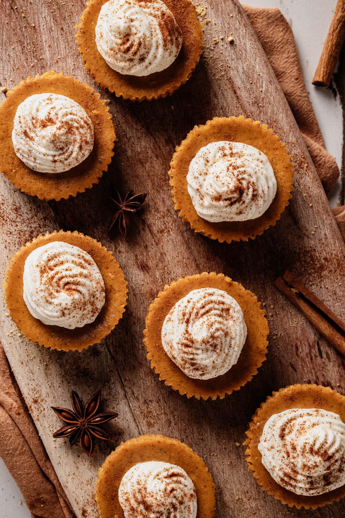 Overhead view of mini pumpkin cheesecakes on a wooden board, each topped with a swirl of whipped cream and dusted with cinnamon.