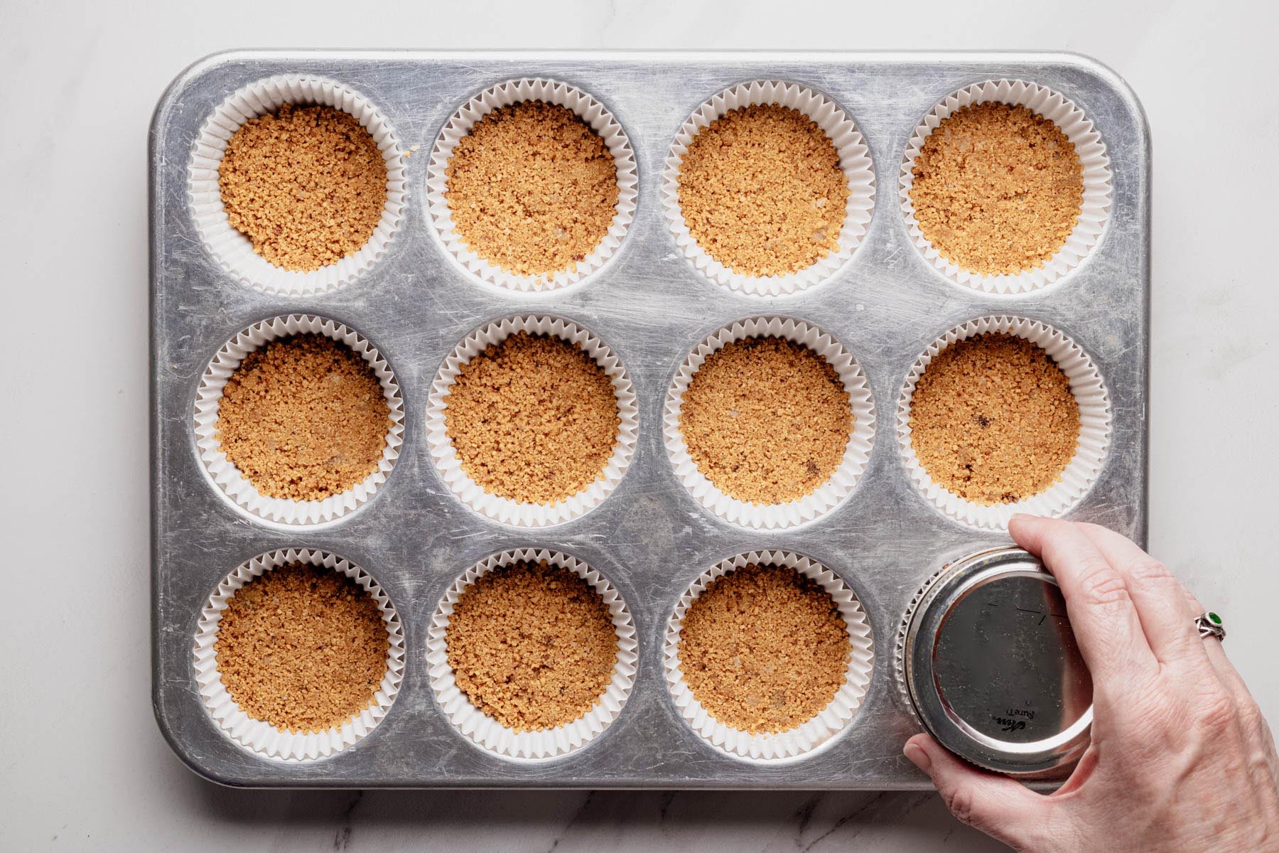 Muffin tin lined with paper cups filled with graham cracker crust, being pressed down with the bottom of a small glass jar.