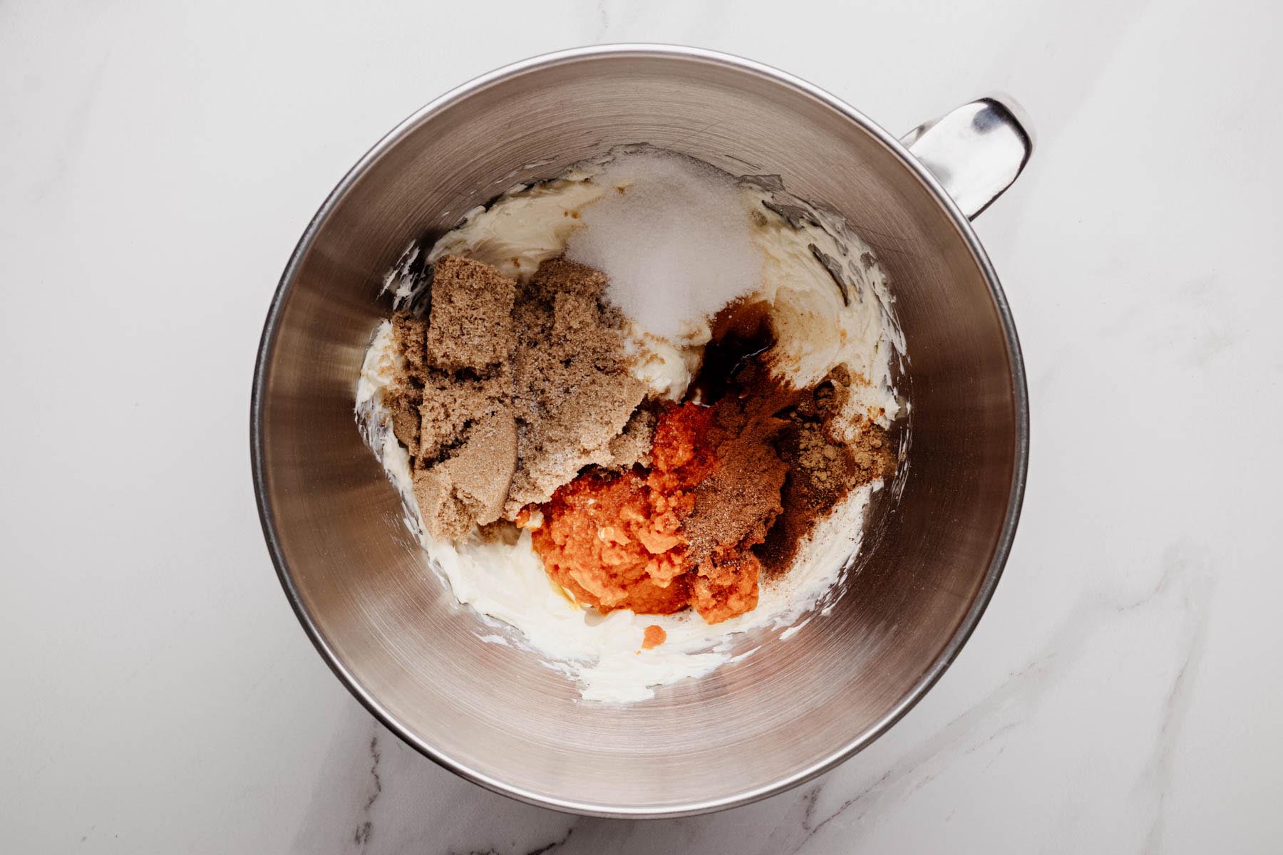 Mixing bowl with cream cheese, pumpkin purée, brown sugar, granulated sugar, vanilla, and spices before mixing for cheesecake filling.