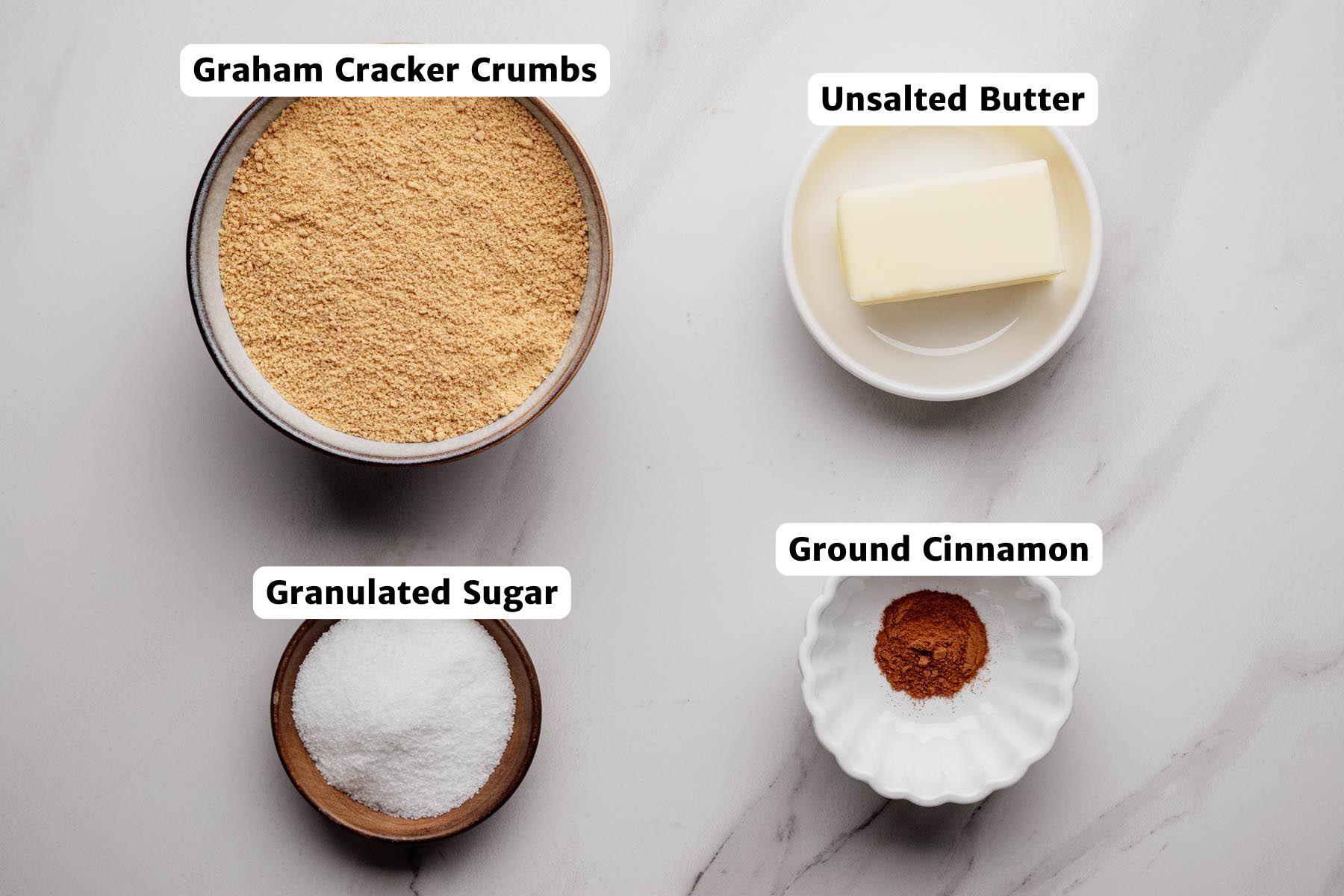 Overhead view of crust ingredients for mini pumpkin cheesecakes, including graham cracker crumbs, granulated sugar, unsalted butter, and ground cinnamon, each labeled in small bowls on a white surface.