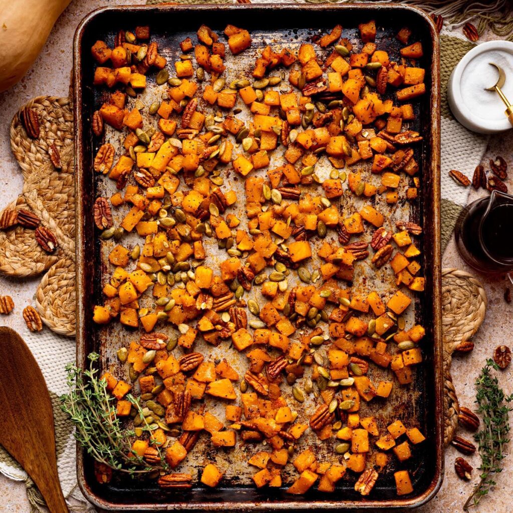 Roasted butternut squash cubes on a sheet pan, caramelized with maple syrup, pecans, and pumpkin seeds, garnished with fresh thyme.
