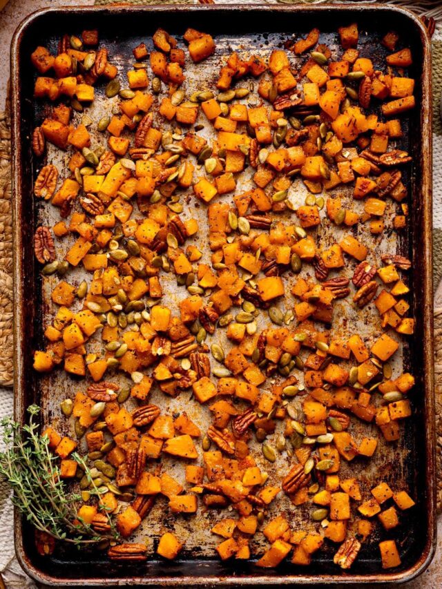 Roasted butternut squash cubes on a sheet pan, caramelized with maple syrup, pecans, and pumpkin seeds, garnished with fresh thyme.
