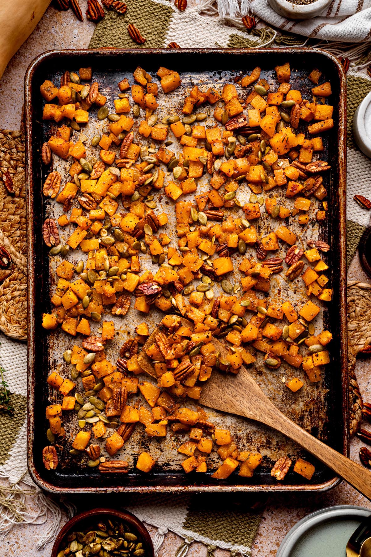 Sheet pan of roasted butternut squash cubes tossed with maple syrup, pecans, and pumpkin seeds, with a wooden spoon for serving.