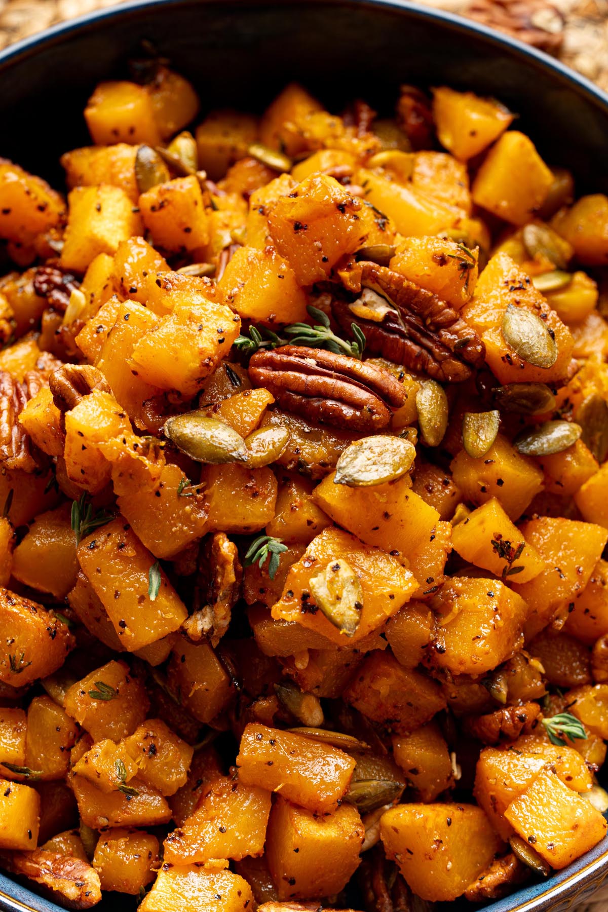 Close-up of roasted butternut squash cubes with maple syrup, pecans, pumpkin seeds, and fresh thyme, served in a dark bowl.