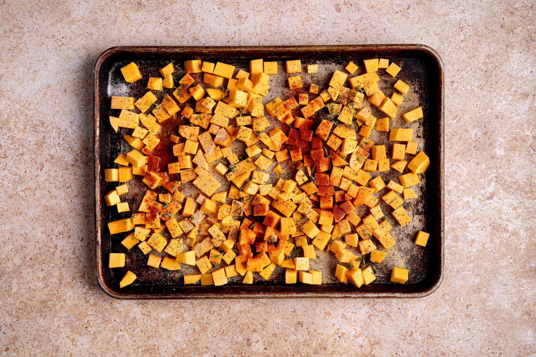 Cubed butternut squash on a sheet pan seasoned with paprika, garlic powder, thyme, salt, and pepper before roasting.