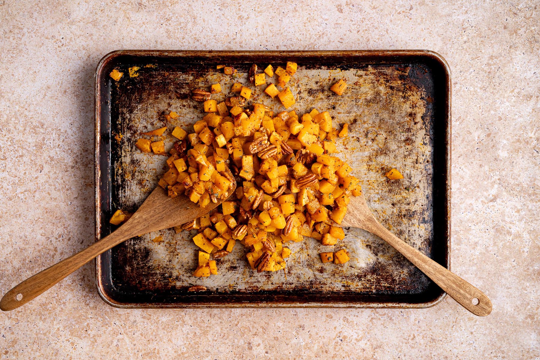 Roasted butternut squash cubes tossed with pecans on a sheet pan using wooden spatulas.