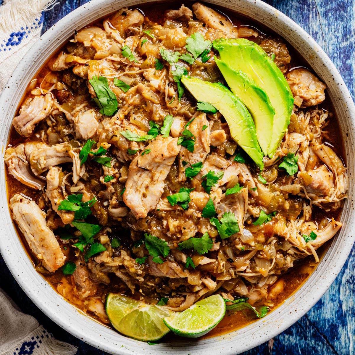 Bowl of shredded green chili chicken topped with avocado slices, fresh cilantro, and lime wedges.