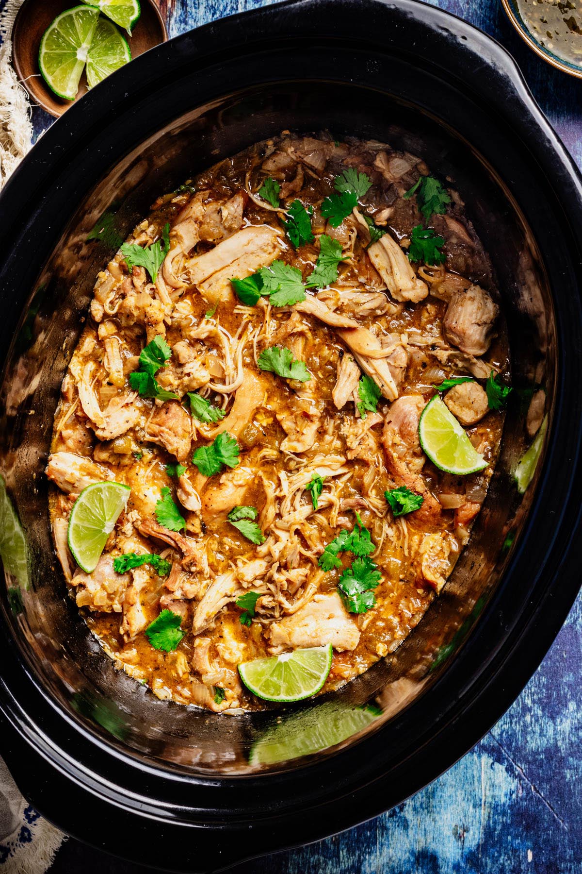 Slow cooker filled with shredded green chili chicken, garnished with cilantro and lime wedges.
