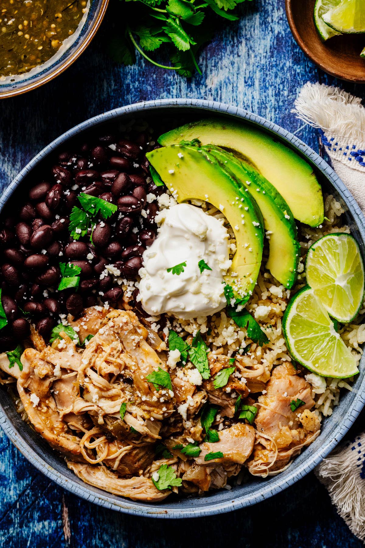 Burrito bowl with slow cooker green chili chicken, black beans, rice, avocado slices, sour cream, cilantro, queso fresco, and lime wedges.