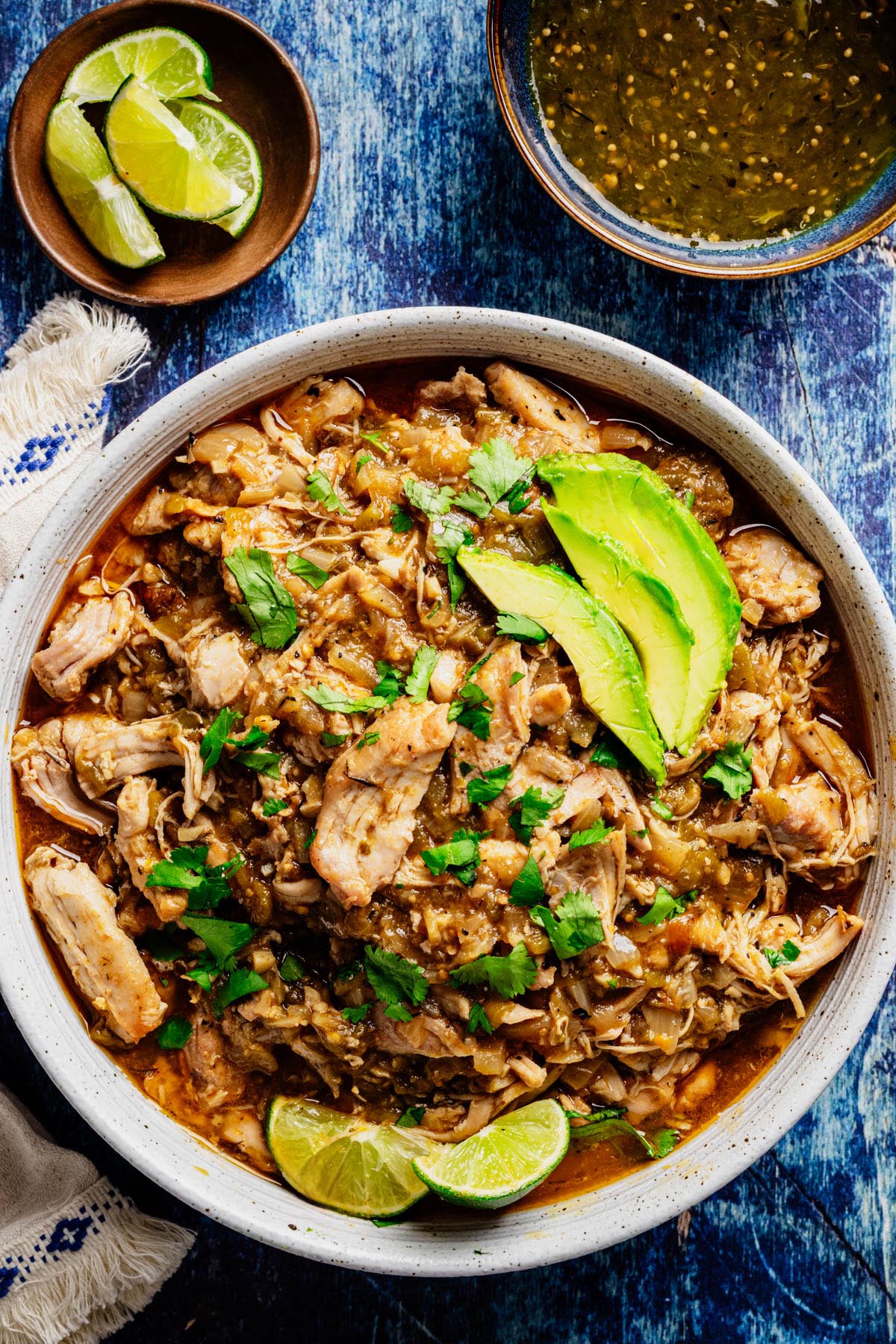 Bowl of slow cooker green chili chicken topped with avocado slices, fresh cilantro, and lime wedges, with salsa verde and extra lime on the side.