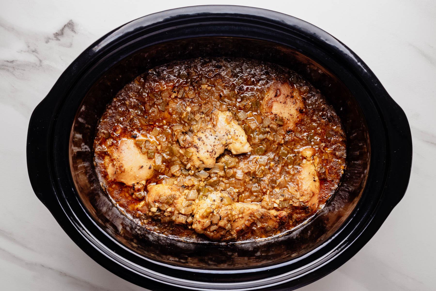 Cooked chicken thighs simmering in a slow cooker with green chili and onion sauce.