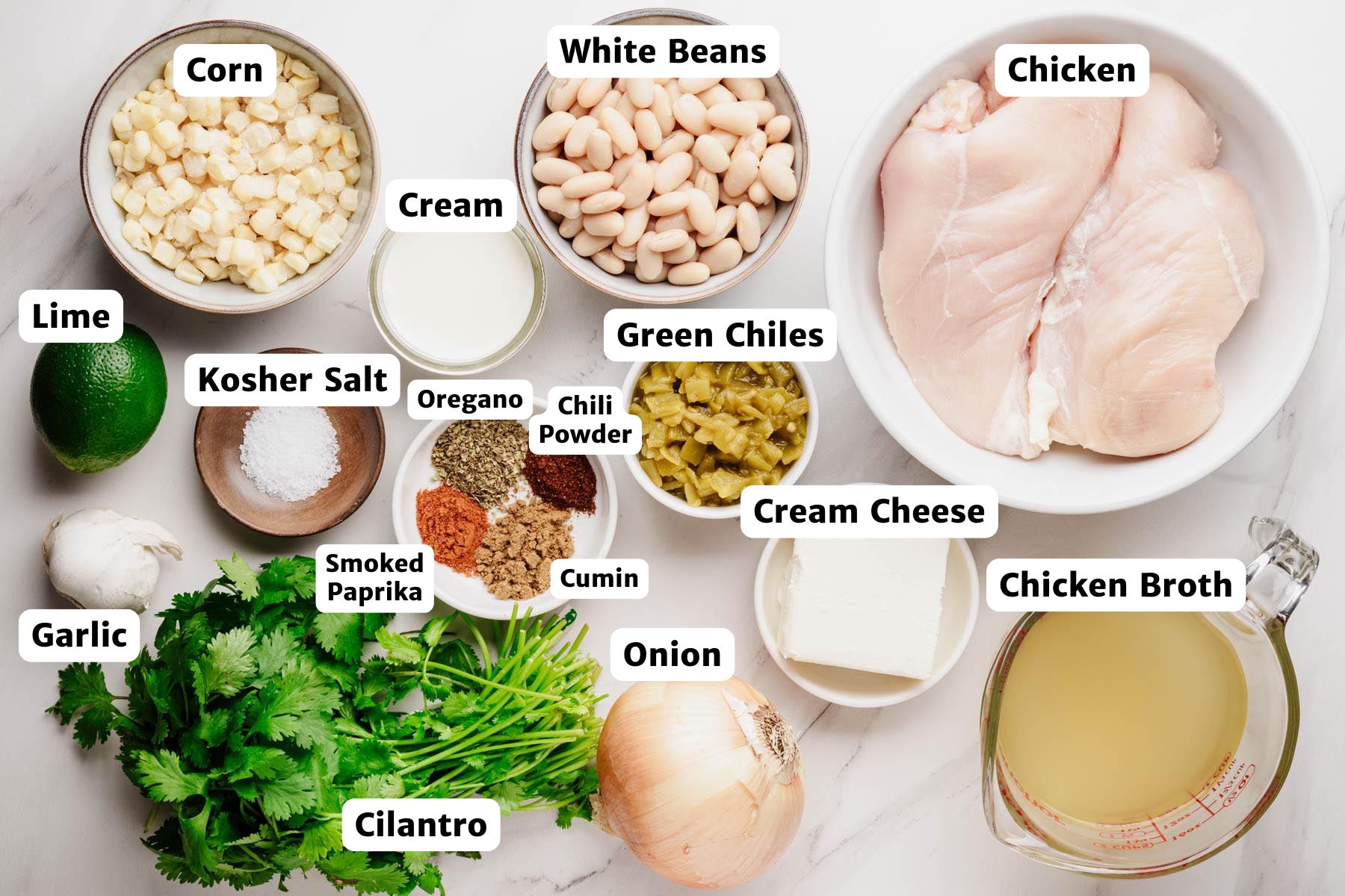 Overhead, labeled ingredients for slow cooker white chicken chili: raw chicken breasts, white beans, corn, green chiles, onion, garlic, cilantro, lime, cream cheese, chicken broth, cream, kosher salt, and spices (cumin, chili powder, smoked paprika, oregano).