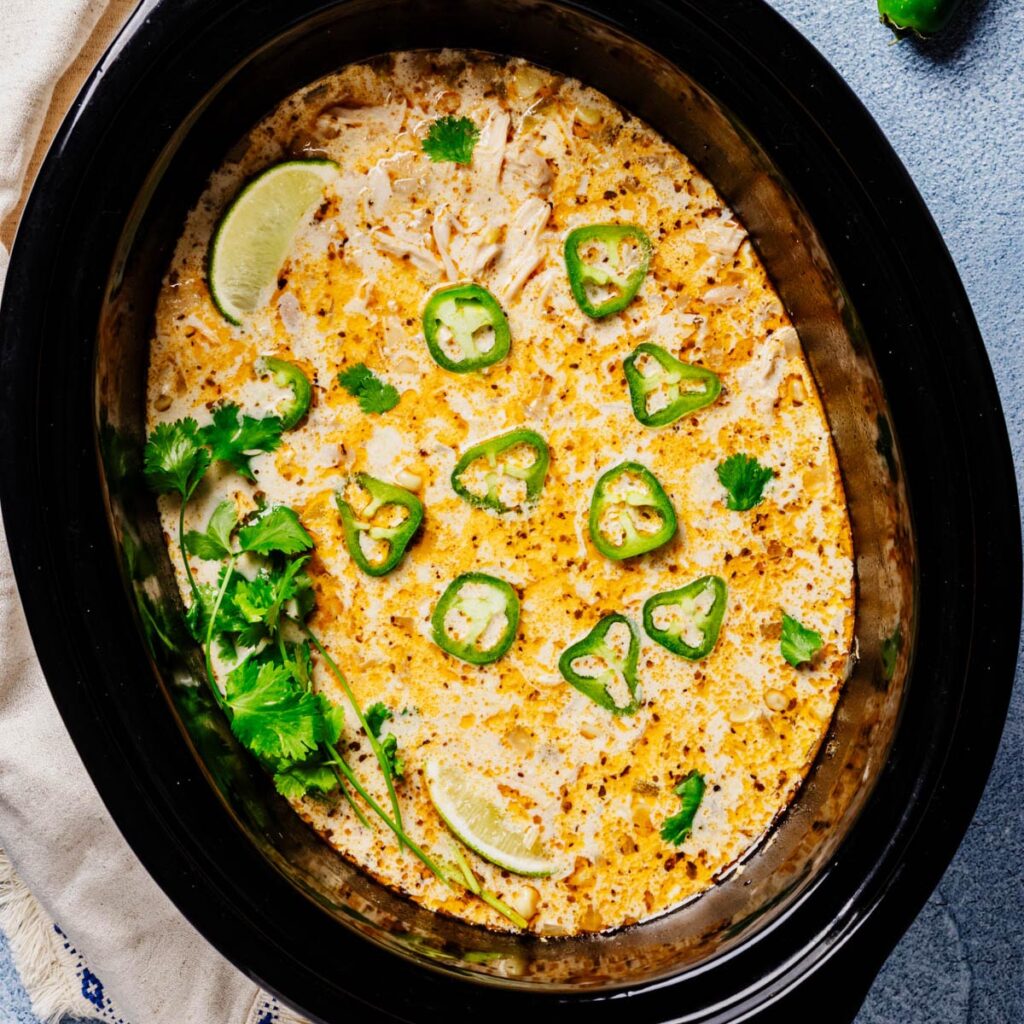 Overhead of a black slow cooker filled with creamy white chicken chili, speckled with spices and topped with jalapeño slices, cilantro, and lime wedges.