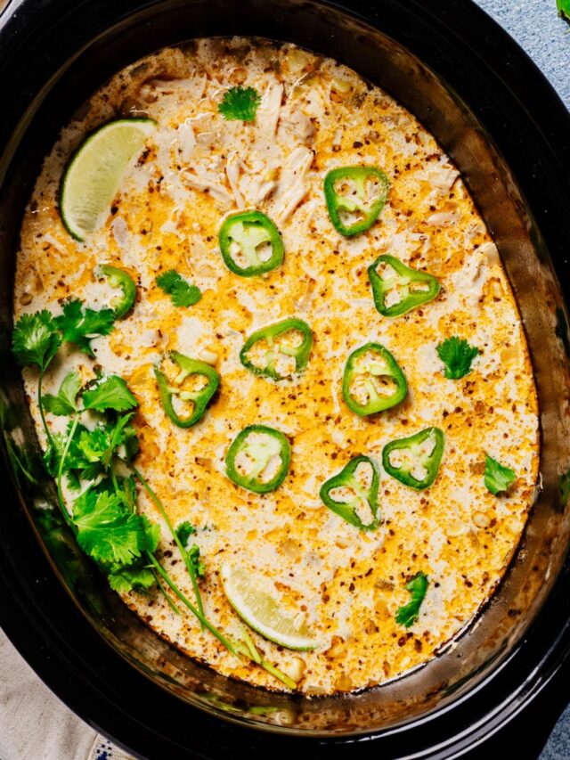 Overhead of a black slow cooker filled with creamy white chicken chili, speckled with spices and topped with jalapeño slices, cilantro, and lime wedges.