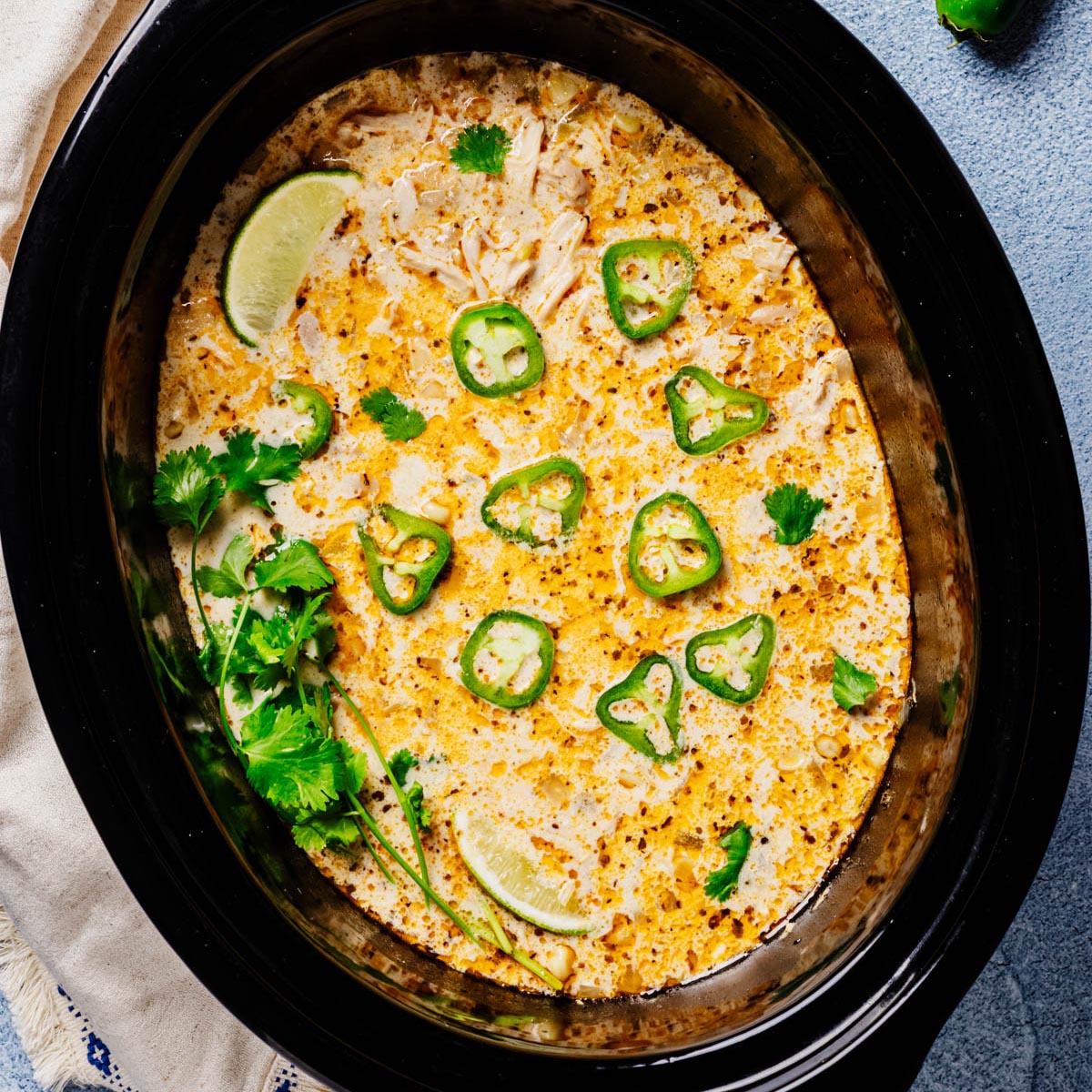 Overhead of a black slow cooker filled with creamy white chicken chili, speckled with spices and topped with jalapeño slices, cilantro, and lime wedges.