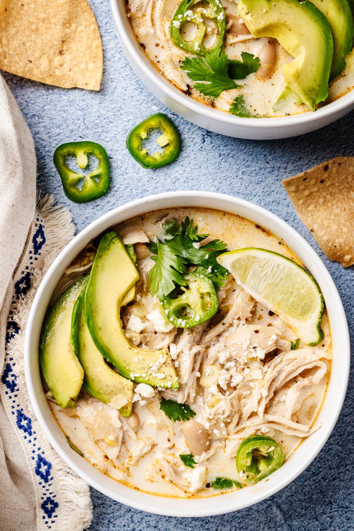 Two bowls of creamy white chicken chili topped with avocado slices, jalapeño, cilantro, and lime, with tortilla chips scattered on a blue surface.