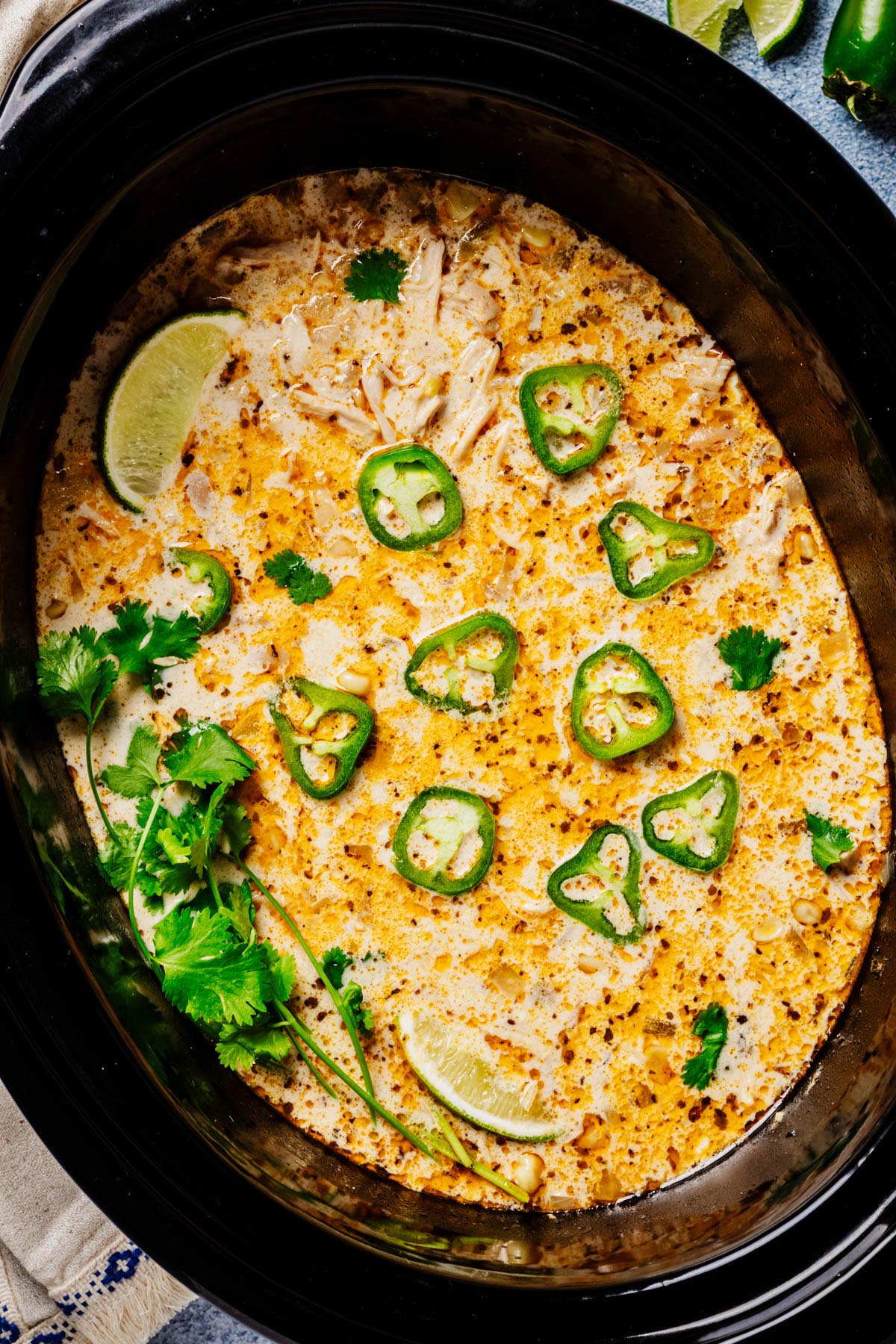 Overhead of a black slow cooker filled with creamy white chicken chili, speckled with spices and topped with sliced jalapeños, cilantro sprigs, and lime wedges.
