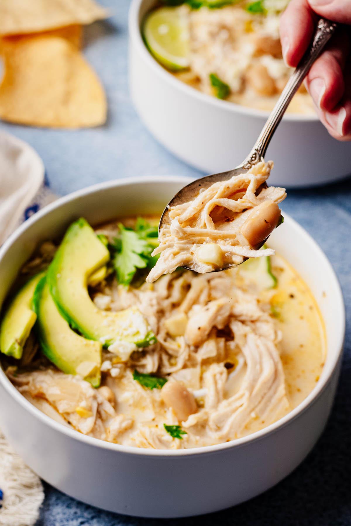 Creamy white chicken chili in a white bowl with shredded chicken, white beans, and cilantro, topped with avocado slices; a spoon lifts a bite above the bowl.