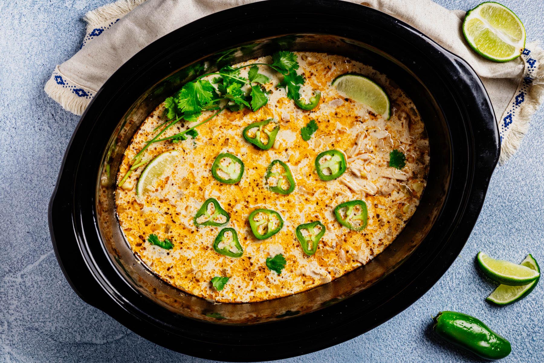 Overhead view of a black slow cooker filled with creamy white chicken chili, topped with jalapeño slices, cilantro, and lime wedges on a blue surface with extra limes.