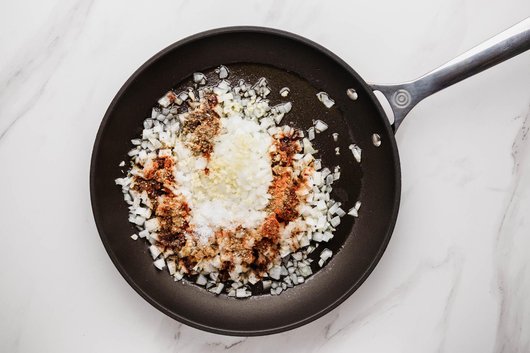 Onions, garlic, and spices (cumin, paprika, oregano, chili powder, salt) blooming in olive oil in a skillet on a white marble surface.