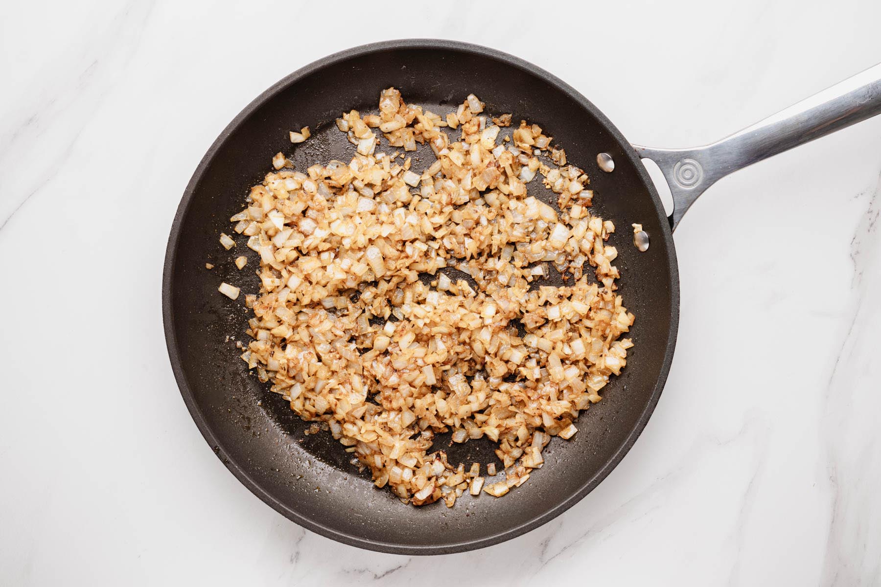 Sautéed diced onions bloomed with spices, lightly browned in a nonstick skillet on a white marble surface.