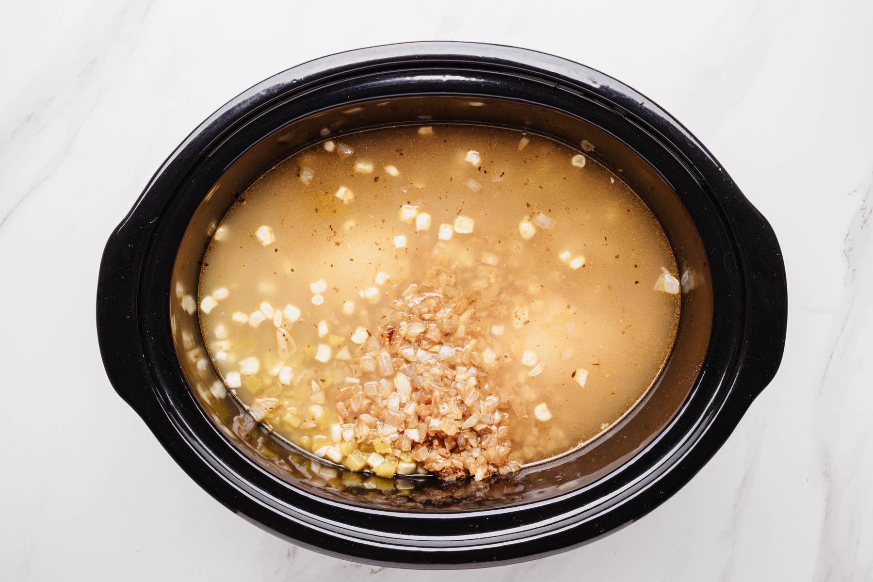 Slow cooker filled with chicken broth, sautéed onions, white beans, green chiles, and corn. The aromatics just added to the crock before cooking.