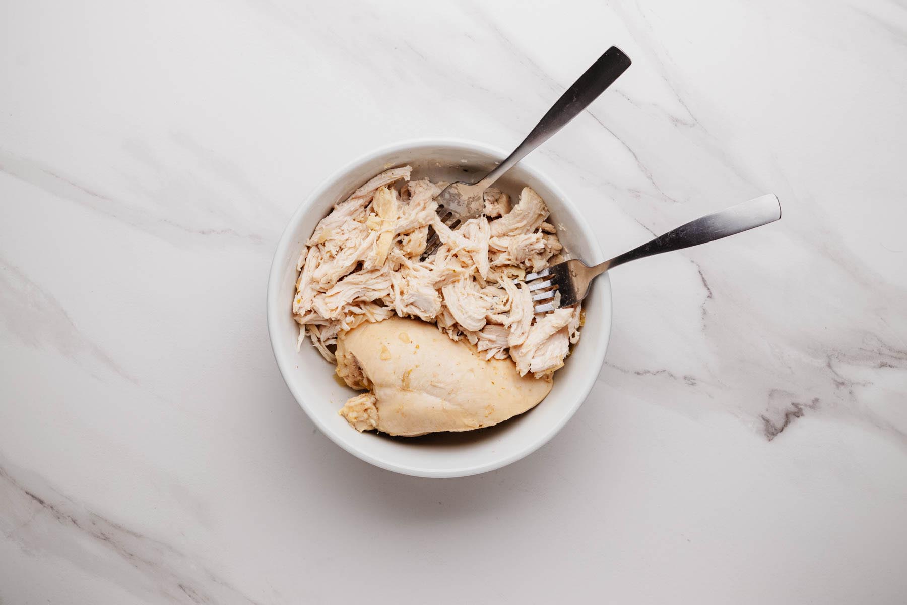 Cooked chicken breast being shredded with two forks in a white bowl on a marble surface.