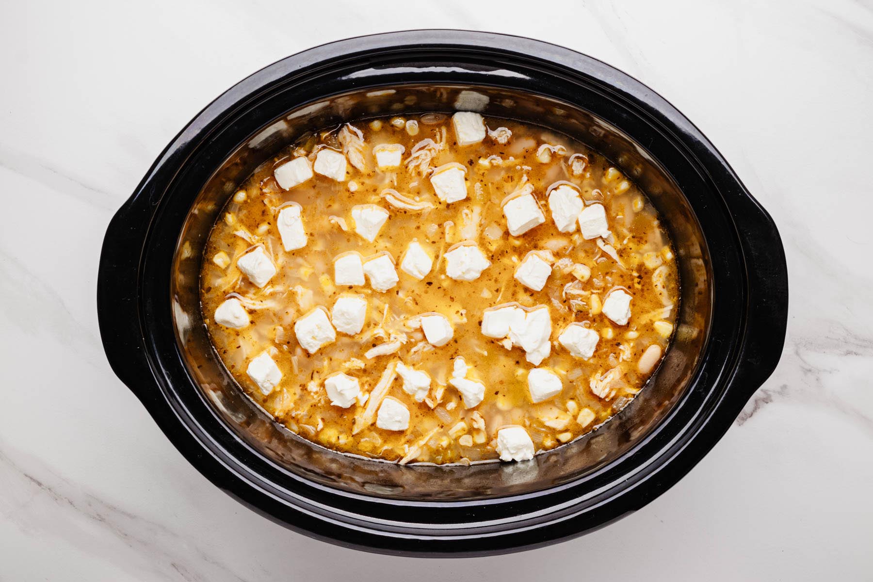 Cubed cream cheese scattered over shredded chicken, beans, and corn in the slow cooker.