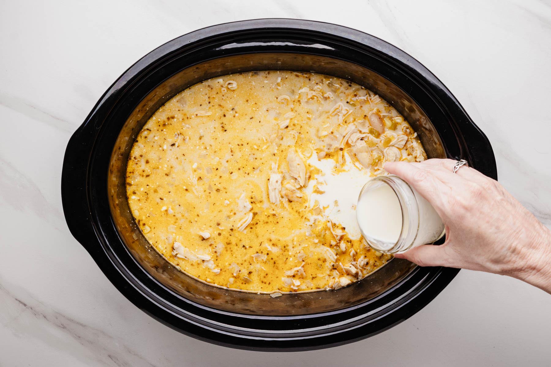 Cream or half-and-half being poured into a slow cooker of white chicken chili after the cream cheese has melted.