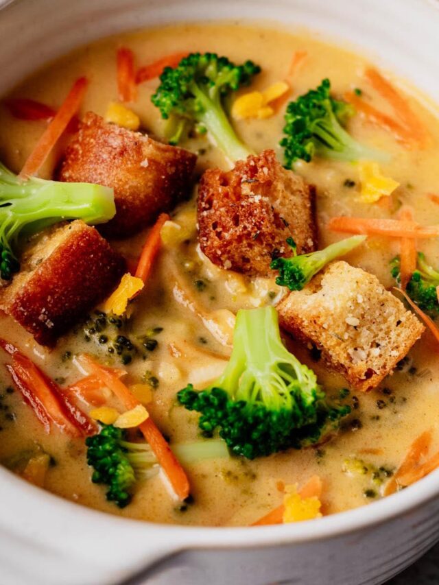 Close-up of creamy broccoli cheddar soup topped with golden croutons, bright broccoli florets, and carrot shreds in a white bowl.