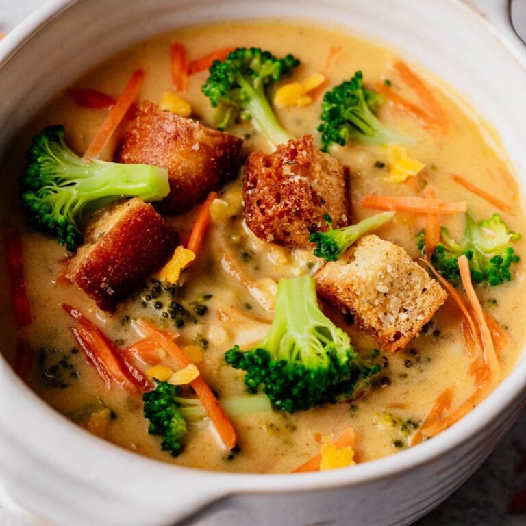 Close-up of creamy broccoli cheddar soup topped with golden croutons, bright broccoli florets, and carrot shreds in a white bowl.