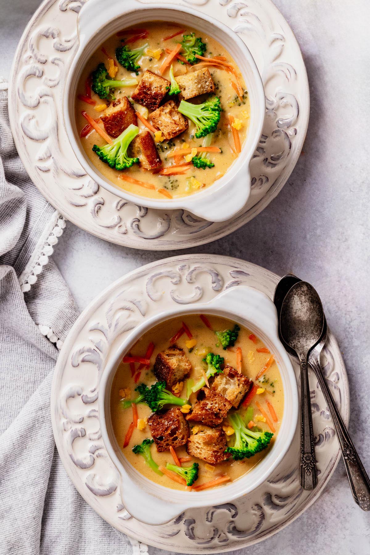 Overhead view of two white bowls of creamy broccoli cheddar soup topped with croutons, broccoli florets, and carrot shreds on ornate plates, with spoons and a gray napkin.