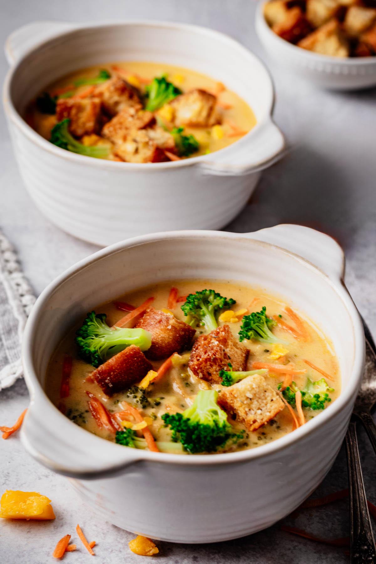 Two white bowls of broccoli cheddar soup, front bowl in sharp focus with golden croutons, broccoli florets, and carrot shreds; second bowl and crouton dish softly blurred in the background.