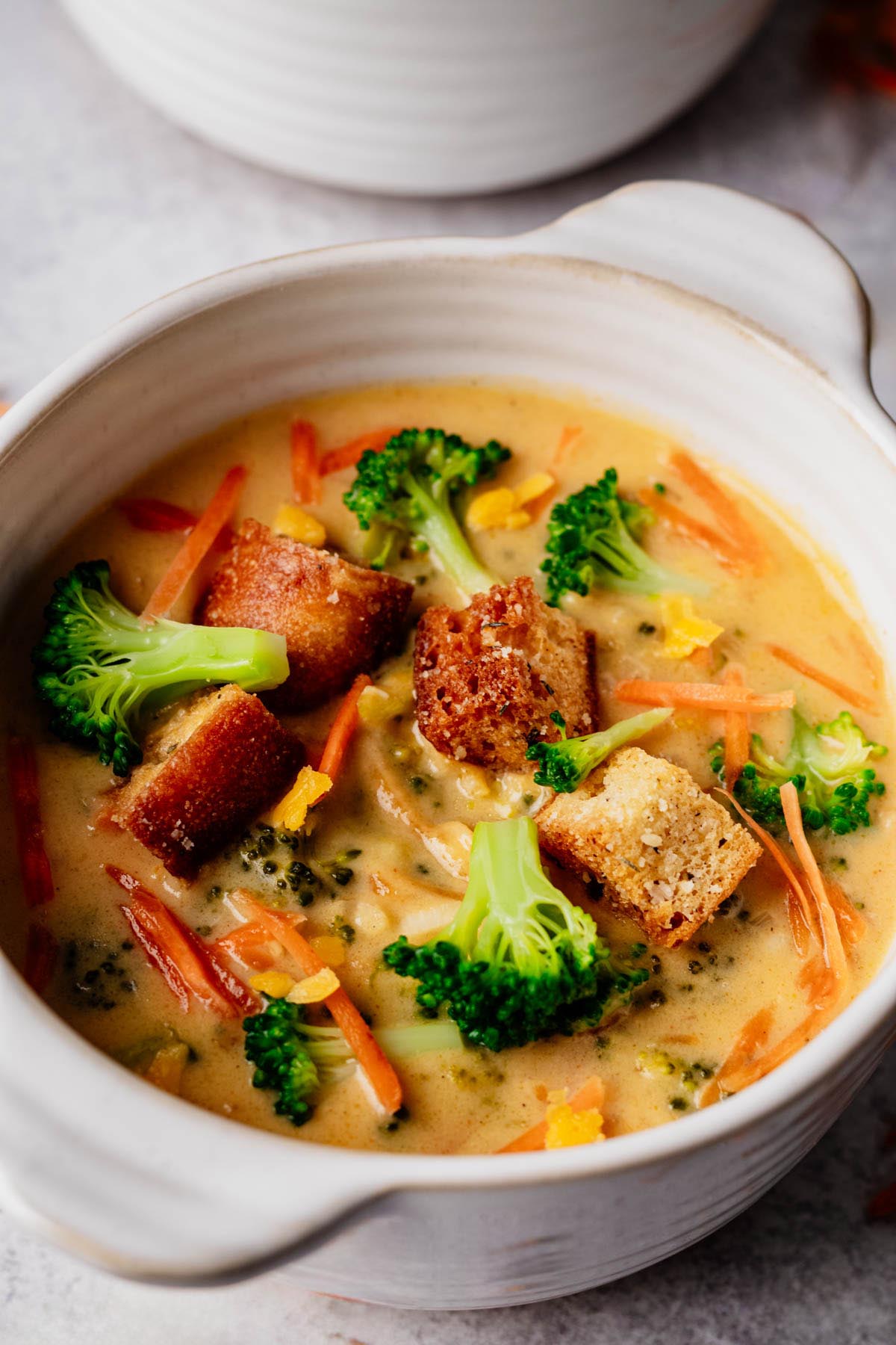 Close-up of creamy broccoli cheddar soup with crisp croutons, tender broccoli florets, and thin carrot strips in a white handled bowl.