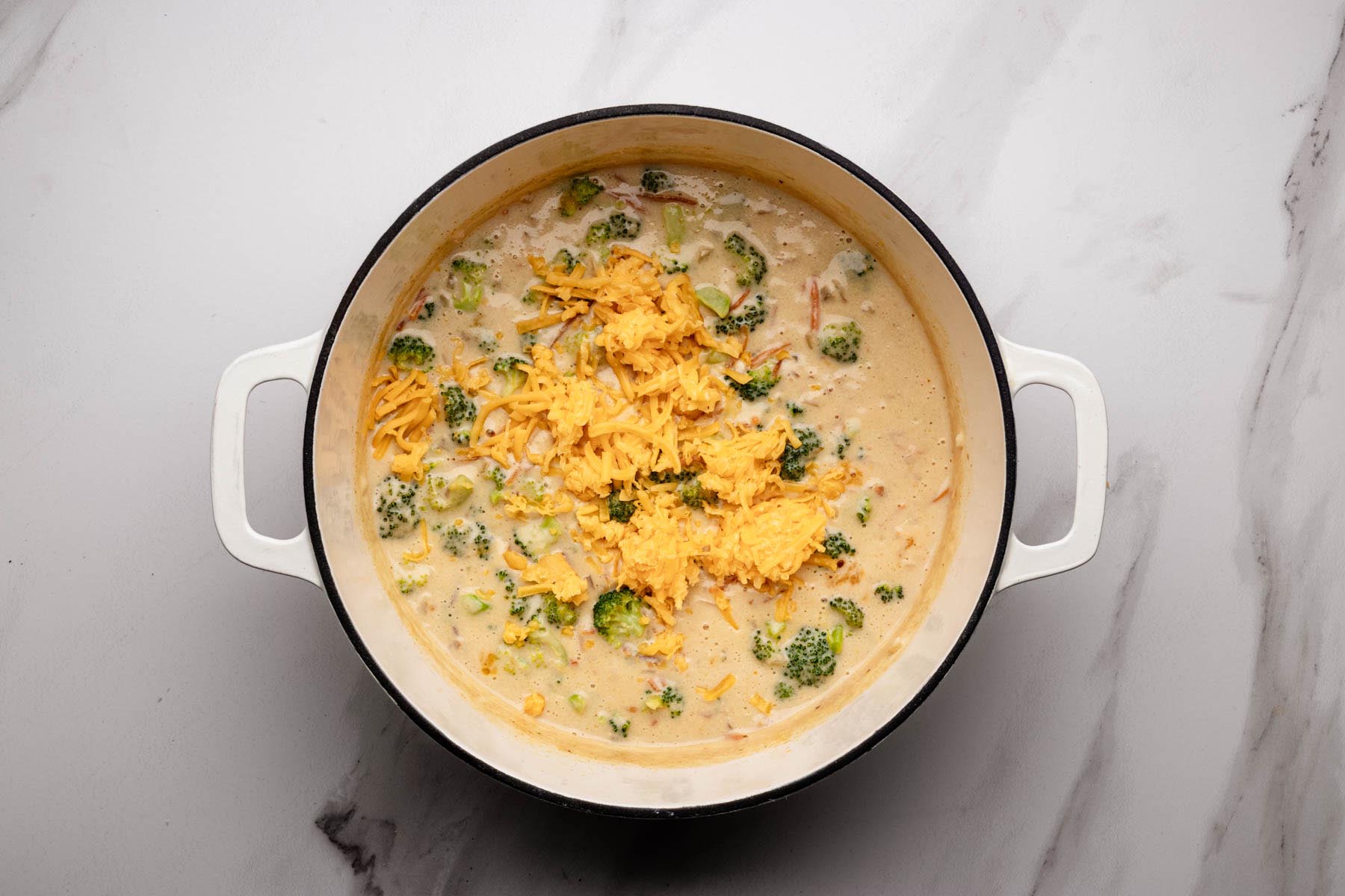 Low-heat pot of creamy broccoli soup with freshly shredded cheddar added in small handfuls to melt.
