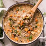 Overhead of a Dutch oven filled with creamy chicken and wild rice soup—carrots, celery, mushrooms, and parsley—with a wooden spoon lifting a scoop.